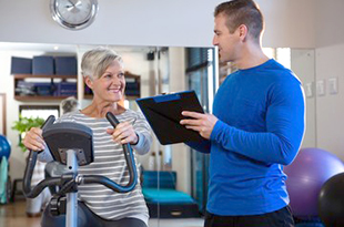 A female patient performs cardiac rehab on a stationary bike while a male physican looks on.
