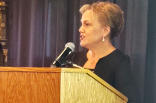 Carol Miller stands behind a podium. She has light brown hair and is wearing drop earrings and a black top.