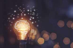 A lightbulb with an illustration of a lit-up brain inside is in front of a dark background with bokeh. An illustration of a network is above the lightbulb. 