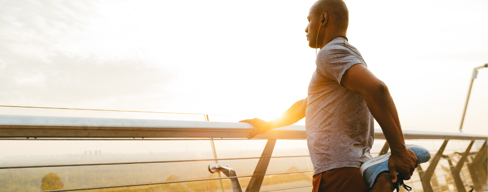 A Black man stretches before a run while peering out at a sunset on a beach.