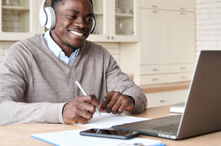 A man sits at a desk watching a zoom meeting on a laptop. He is smiling and wearing white headphones.