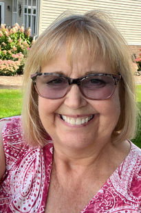 A portrait of Crystal Mroz. A woman with blonde hair, crystal is wearing a pink and white patterned top and a pair of glasses.