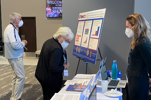 BMC2 Project Manager, Annemarie Forrest, greets summit attendees at the BMC2 table. There are a variety of pamphlets on the table and a large poster display to the left of the table. A man and a woman look on.