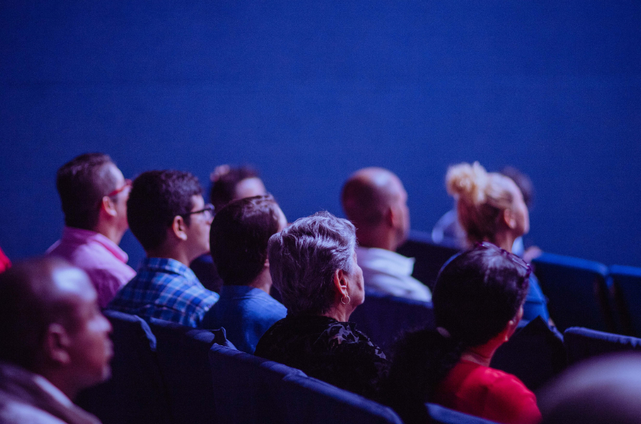 A view of an audience from the back right. The room is low-lit and has a blue cast.