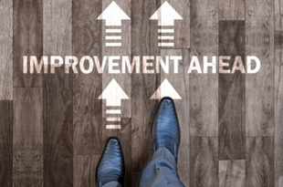 In this view from above, a man's feet step toward arrows facing forward and the words "improvement ahead" painted on the wooden floor.