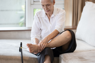 A man wearing a white shirt and black pants sits on the edge of a bed rubbing his ankle. The bed has a white pillow case and comforter and there is a black cane resting on it to the man's right.