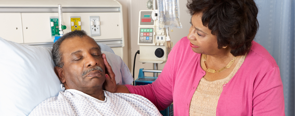 A Black patient lies in a hospital bed with his eyes closed while his wife, dressed in pink, strokes his cheek.