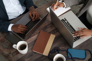 In a view from above, cropped so you cannot see their faces, a Black man and Black woman in business attire sit at a wooden table working on their laptops. Two white coffee mugs, a book, a phone, and headphones are also on the table.