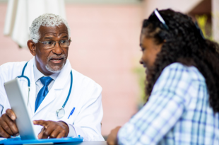 A Black doctor reviews resources on an iPad with a patient. He has short grey hair and is wearing glasses, a light colored shirt, a white coat, and blue tie and stethoscope. He also has a blue pen in his pocket. The patient is a Black woman with long curly hair. She has eyeglasses on her head and is wearing a long-sleeved white and blue plaid shirt.