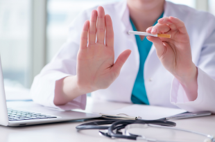 A female physician, wearing blue scrubs under a white coat, sits at her desk surrounded by a laptop, stethoscope, and papers. She is holding a cigarette in her left hand and making a "stop" signal with the other. Her face is not seen.