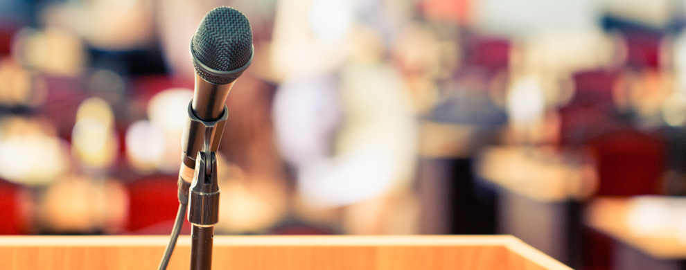 A microphone at a podium is photographed using shallow depth of field with an audience out of focus in the background.
