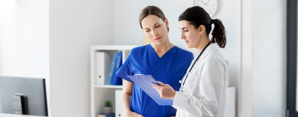 Two female health care professionals, one in blue scrubs the other in a white coat, review a patient chart on a clip board in an office setting.