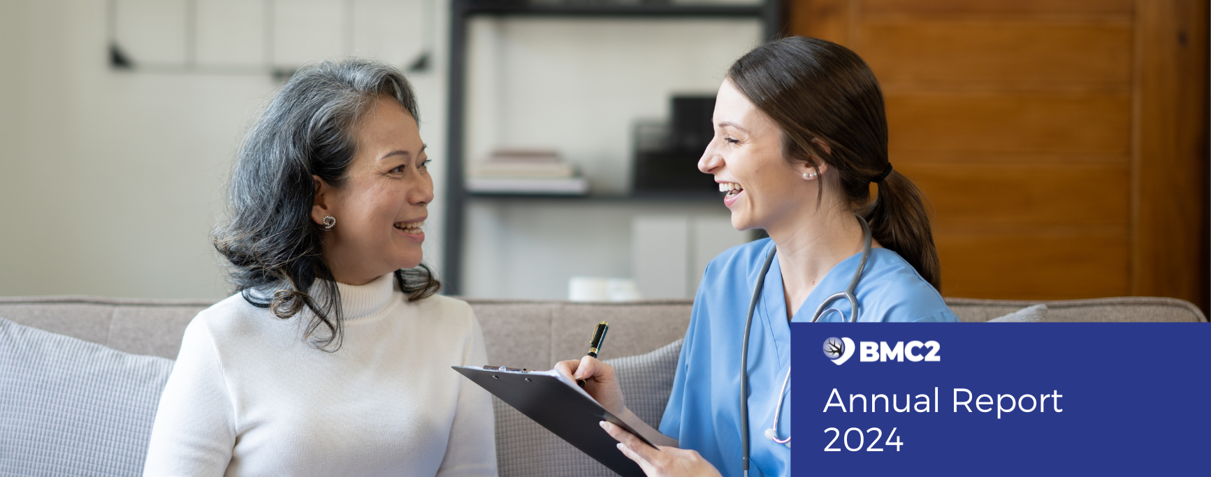 A young white health care provider smalls while speaking with a patient and writing notes on a clip board. She is smiling, wears blue scrubs and a stethoscope around her neck and has long dark hair pulled back in a pony tail. Her patient, an older Asian woman with shoulder-length salt and pepper hair, wearing a white turtle neck sweater, smiles back.