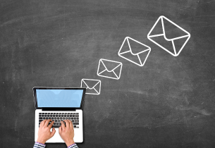 A view from above of a man's hands typing on a laptop keyboard sitting on a chalkboard table.  envelopes in progressily larger sizes are drawn in white chalk from the upper right hand corner of the laptop screen.