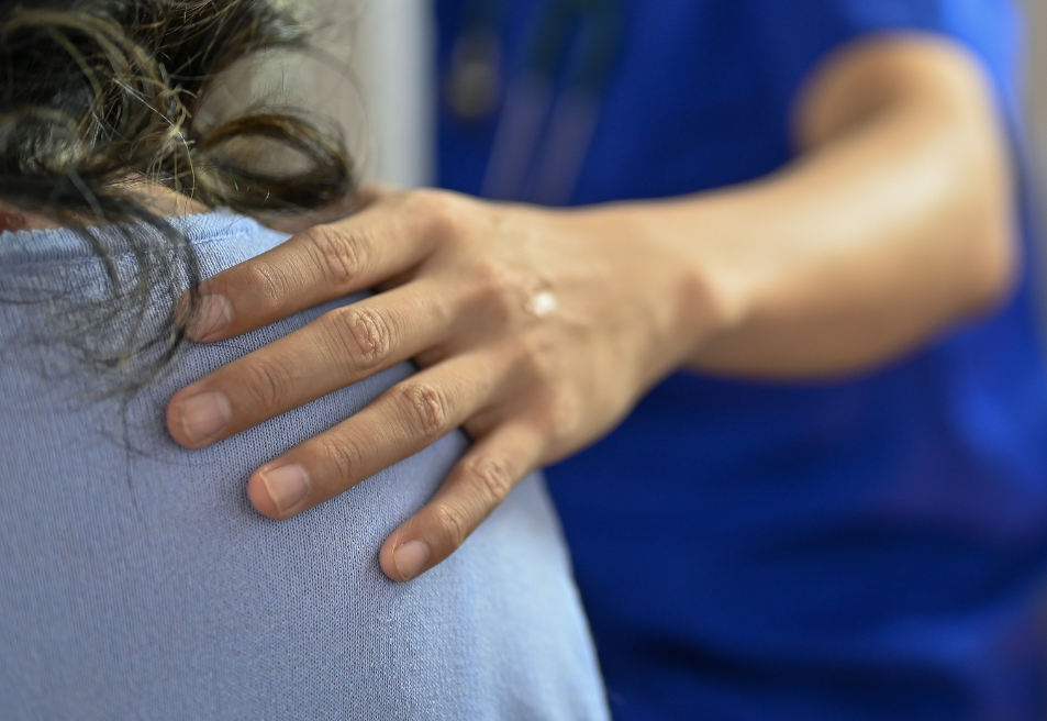A health care provider wearing bright blue scrubs reaches out to put a hand on the shoulder of a colleague wearing a light blue top.