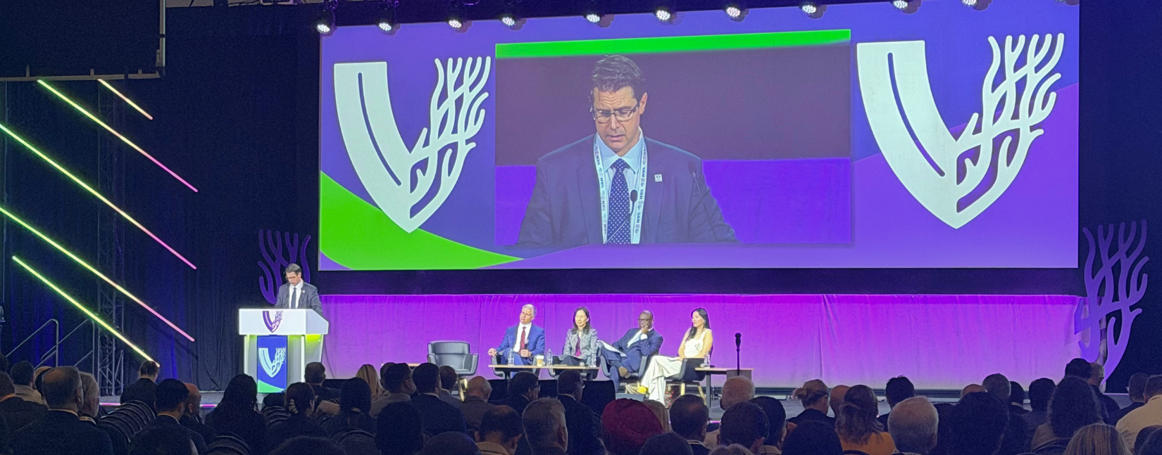 Dr. Osborne stands at the podium on the #VAM2026 stage. He's wearing a blue suit and his image is projected on a giant screen behind him.