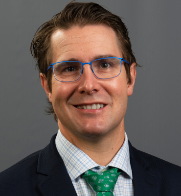 Dr. Nick Osborne has short brown hair and blue-rimmed glasses. He is smiling and wearing a dark jacket, light-colored windowpane patterned collared dress shirt, and a green tie.