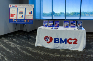 BMC2's display at the 2022 Obesity Management Summit. A table displays a variety of hand outs about BMC2 and there is a large poster with BMC2 data to the left.